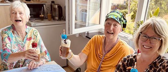 Drei Frauen sitzen an einem Tisch mit Getränken und Blumen. Sie lächeln und scheinen eine Mahlzeit zu genießen. Eine Mikrowelle und eine Kaffeemaschine sind im Hintergrund zu sehen.
