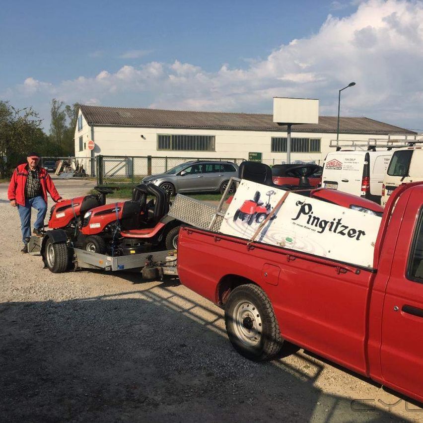 Ein Mann steht neben einem roten Pickup-Truck, der einen Rasenmäher zieht. Der Truck hat ein Schild mit der Aufschrift 'Pingitzer' auf dem Laderaum. Hinter den Fahrzeugen befindet sich ein großes weißes Gebäude.