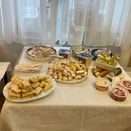 Ein Tisch mit einer weißen Tischdecke ist mit einer Auswahl an Lebensmitteln beladen, darunter Gebäck, Brot, Joghurt und Käse, mit einem Fenster mit weißen Vorhängen im Hintergrund.