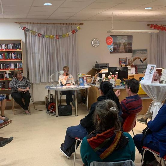 In einer Bibliothek sitzt eine Frau an einem Tisch und hält eine Rede, während andere zuhören.