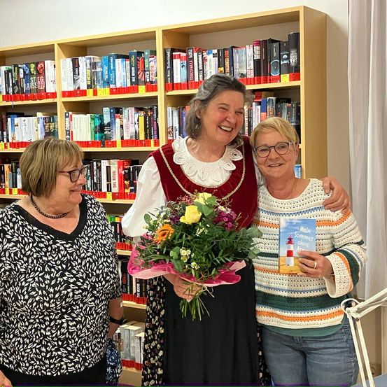 Drei lächelnde Frauen stehen vor einem mit Büchern gefüllten Bücherregal. Die Frau in der Mitte hält einen Blumenstrauß und ein Buch mit einem Leuchtturm auf dem Cover.