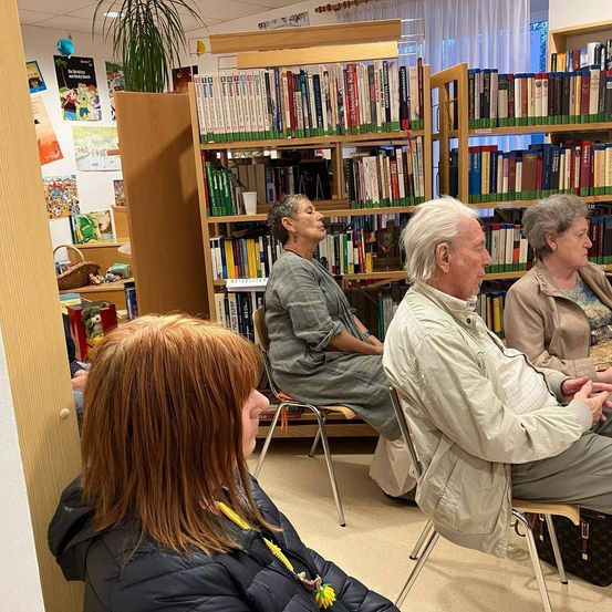 Drei ältere Erwachsene sitzen im Kreis und sehen sich an. Eine Frau mit einer Halskette sitzt neben einem Mann in einer Jacke. Hinter ihnen sitzt eine Frau auf einem Stuhl, und ein Mann steht neben einem Bücherregal, das mit Büchern gefüllt ist.