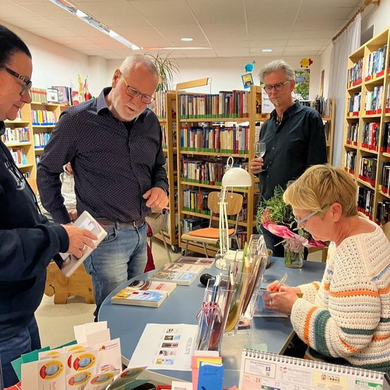 Drei Männer und eine Frau sind in einer Buchhandlung. Ein Mann schreibt in ein Notizbuch, während die anderen stehen und reden. Auf dem Tisch liegen Bücher und eine Lampe, und Regale mit Büchern dahinter.
