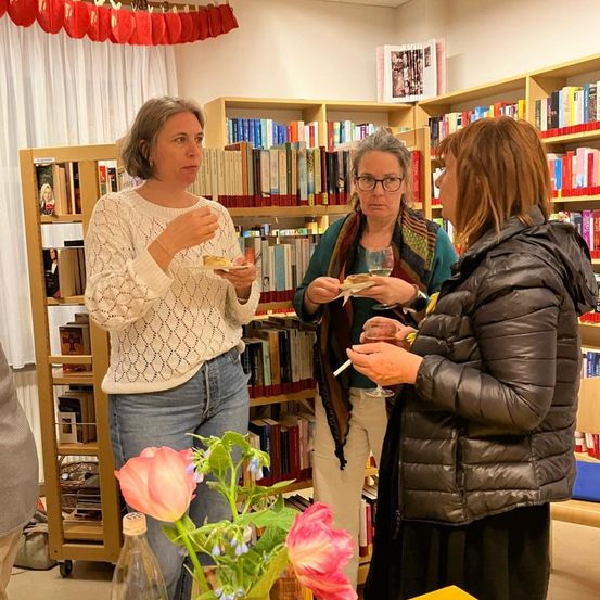 Drei Frauen stehen in einem Raum mit Bücherregalen im Hintergrund. Die Frau auf der linken Seite hält einen kleinen Teller mit Essen und spricht mit der Frau in der Mitte. Die Frau in der Mitte hält ein Weinglas und spricht mit der Frau auf der rechten Seite.