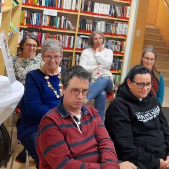 Eine Gruppe von fünf Personen sitzt in einer Buchhandlung. Sie sitzen auf Stühlen und eine Person sitzt auf einer Bank. Sie sehen in die Kamera und lächeln.