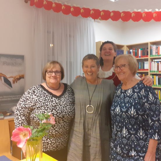 Vier Frauen stehen in einem Raum und lächeln. Zwei von ihnen tragen eine Brille. Eine hat eine Halskette. Dahinter steht ein Bücherregal mit Büchern.