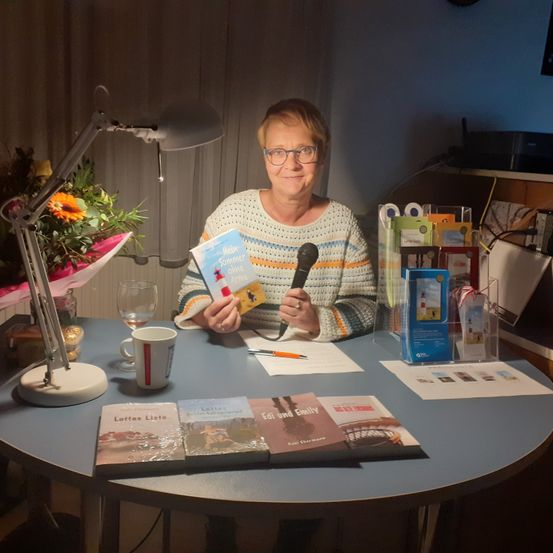 Eine Frau sitzt an einem Tisch mit Büchern, einem Mikrofon und einer Lampe. Sie hält ein Buch mit dem Titel 'Mein Sommer' und lächelt in die Kamera.