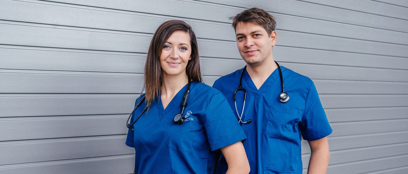 Ein Mann und eine Frau in blauen Scrubs stehen nebeneinander und lächeln für ein Foto. Beide haben Stethoskope um den Hals.