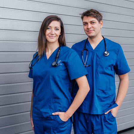 Eine lächelnde Frau und ein Mann, beide in blauen Kitteln, posieren für ein Foto vor einem grauen Hintergrund.