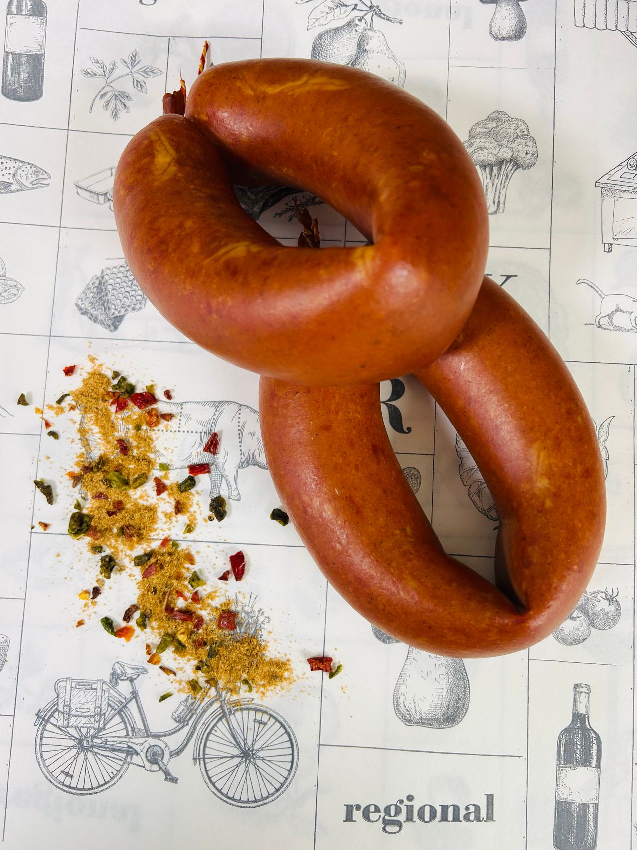 Zwei Würste sind auf einem weißen gemusterten Papier zu einem Knoten gebunden, mit einigen Chiliflocken und Brotkrümeln verstreut. Im Hintergrund gibt es Zeichnungen von einem Fisch, Brokkoli und einer Katze.