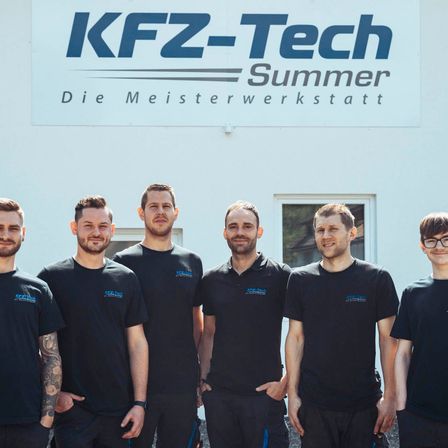 Eine Gruppe von Männern in schwarzen T-Shirts mit blauem Text steht vor einem Gebäude mit einem großen Schild, auf dem KFz-Tech Summer steht. Sie lächeln und scheinen für ein Foto zu posieren.