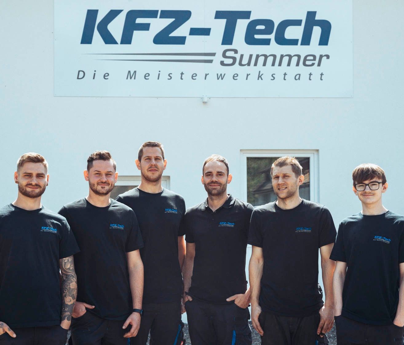 Eine Gruppe von Männern in schwarzen T-Shirts mit blauem Text steht vor einem Gebäude mit einem großen Schild, auf dem KFz-Tech Summer steht. Sie lächeln und scheinen für ein Foto zu posieren.