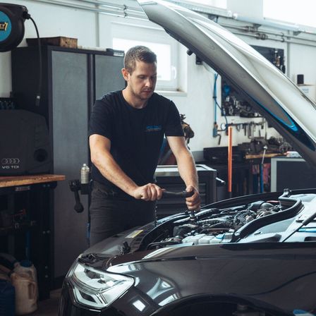 Ein Mann in einer Garage mit geöffneter Motorhaube eines schwarzen Autos, hält eine Schraube.