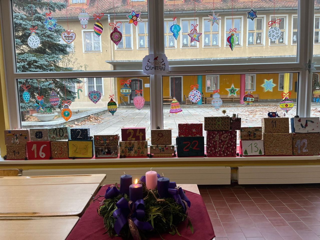 Ein Raum mit nummerierten Boxen von 1 bis 24, dekorativen Ornamenten und einem Tisch mit einem Kranz und Kerzen. Draußen ist ein Blick auf ein Schulgebäude mit Weihnachtsdekorationen an den Fenstern.
