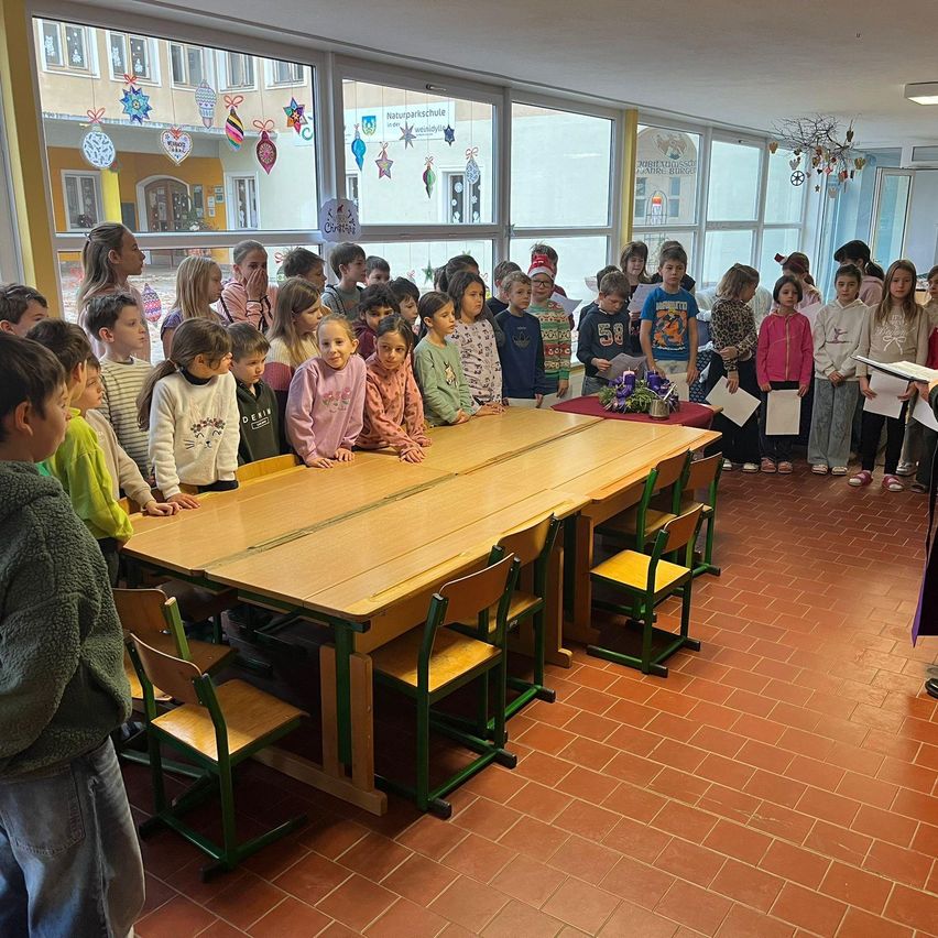 Eine Gruppe von Kindern in einem Klassenzimmer steht und sitzt um einen langen Tisch herum, möglicherweise während einer Zeremonie. Eine Person steht vor dem Tisch mit einem Blumenstrauß.
