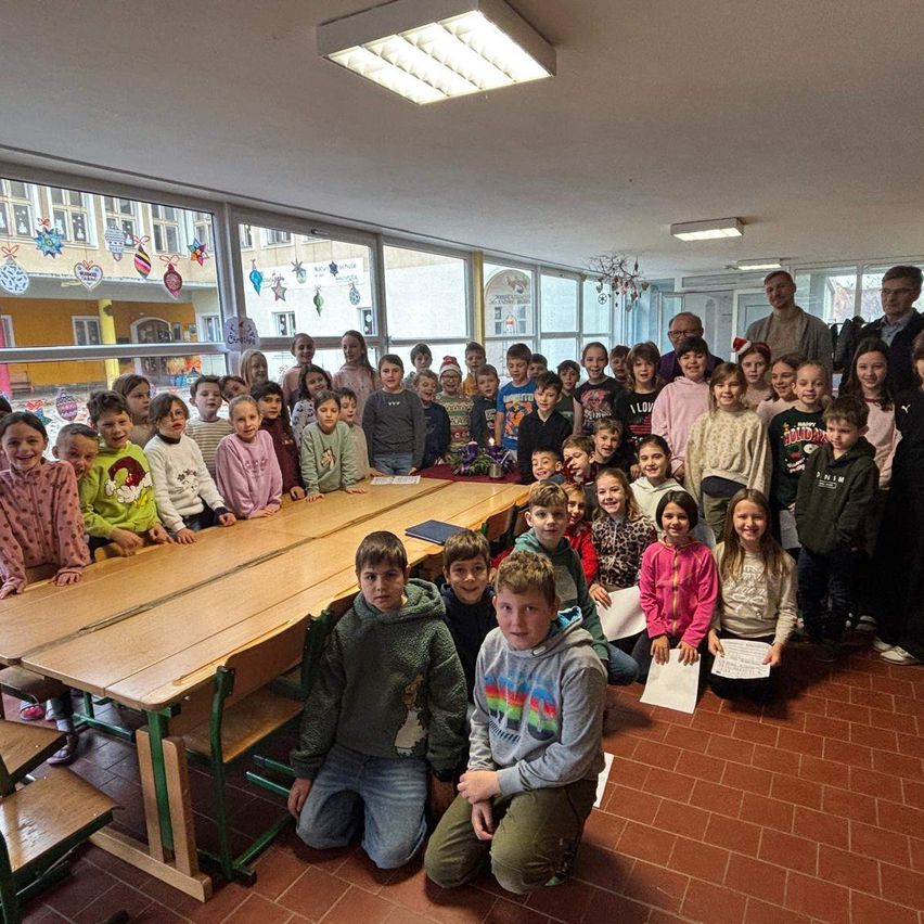 Eine große Gruppe von Kindern und Erwachsenen posiert für ein Foto in einem Klassenzimmer. Einige sitzen auf dem Boden, während andere hinter Tischen stehen. Der Raum hat große Fenster und Dekorationen.