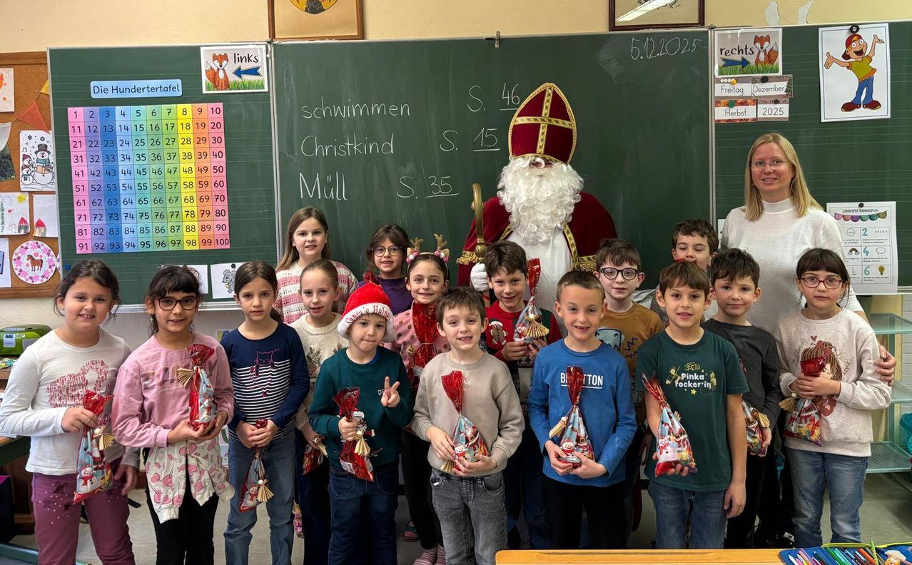 A group of children in a classroom are posing for a photo with a man dressed as Santa Claus. Some of them are holding gifts. Behind them is a chalkboard with numbers and words written on it.