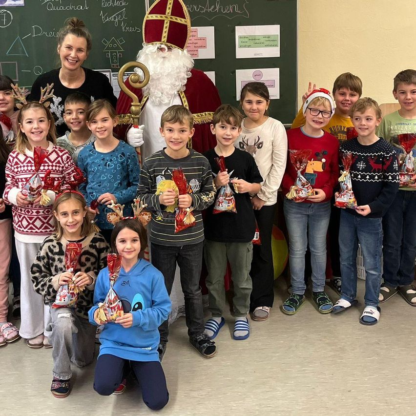 A group of children poses for a photo with Santa Claus, holding gifts in a classroom.