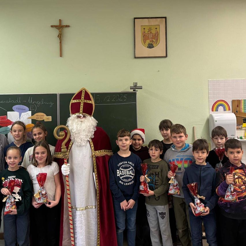 A group of children poses with a person dressed as Santa Claus in a classroom, holding gifts. A blackboard and a cross are in the background.