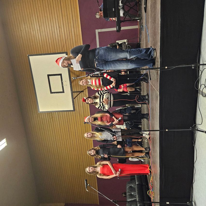A choir, dressed in festive attire, performs on stage with a wooden wall backdrop. A pianist sits in the background.