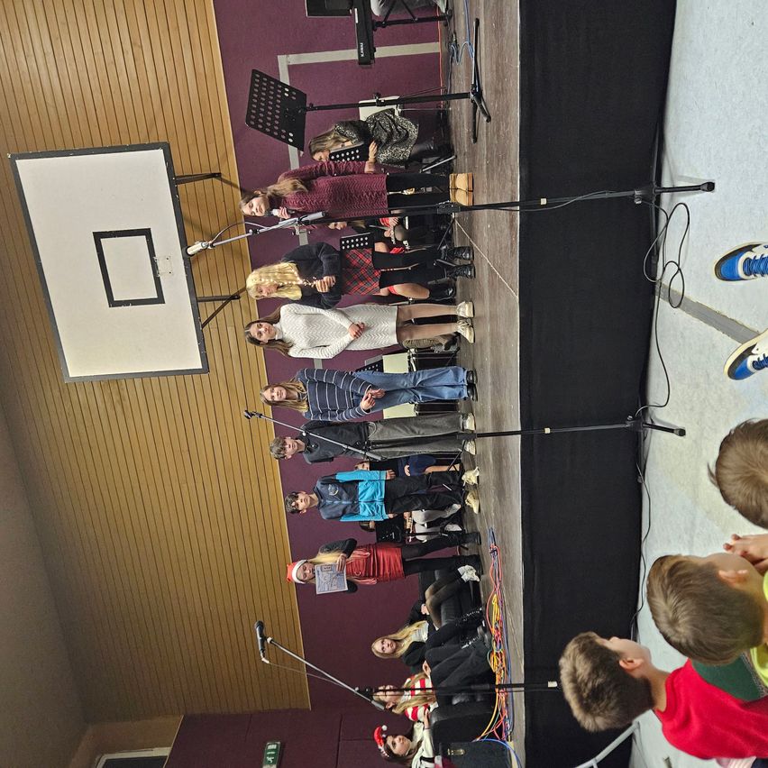 A group of children and adults on a stage with microphones, possibly for a performance. A basketball hoop is on the left. Audience is sitting in front.