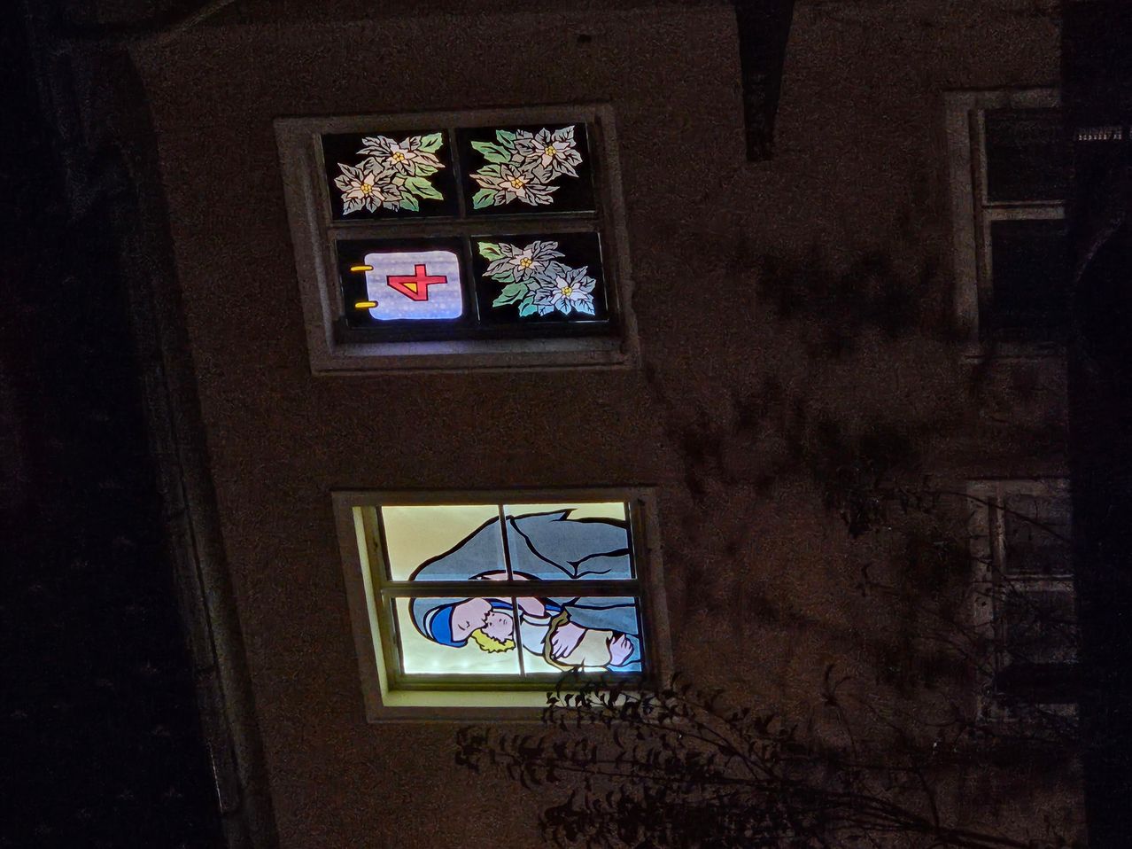 Two illuminated stained glass windows on a building. The top one has a red cross and floral patterns. The bottom one depicts a person carrying a child.