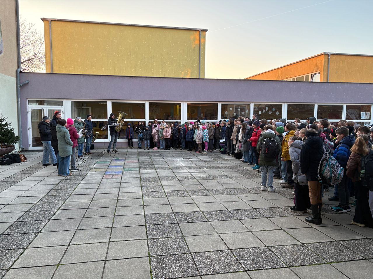 A group of people gather outdoors near a building, with musicians playing instruments in the center. Some are wearing hats and gloves, indicating cold weather. The building has glass windows and a warm glow from lights inside.
