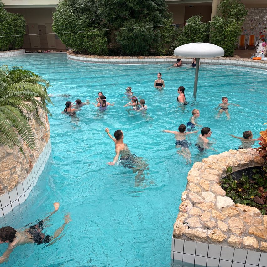 A group of people swim in a large indoor pool, with several wearing goggles. Some swimmers are in the shallow end.