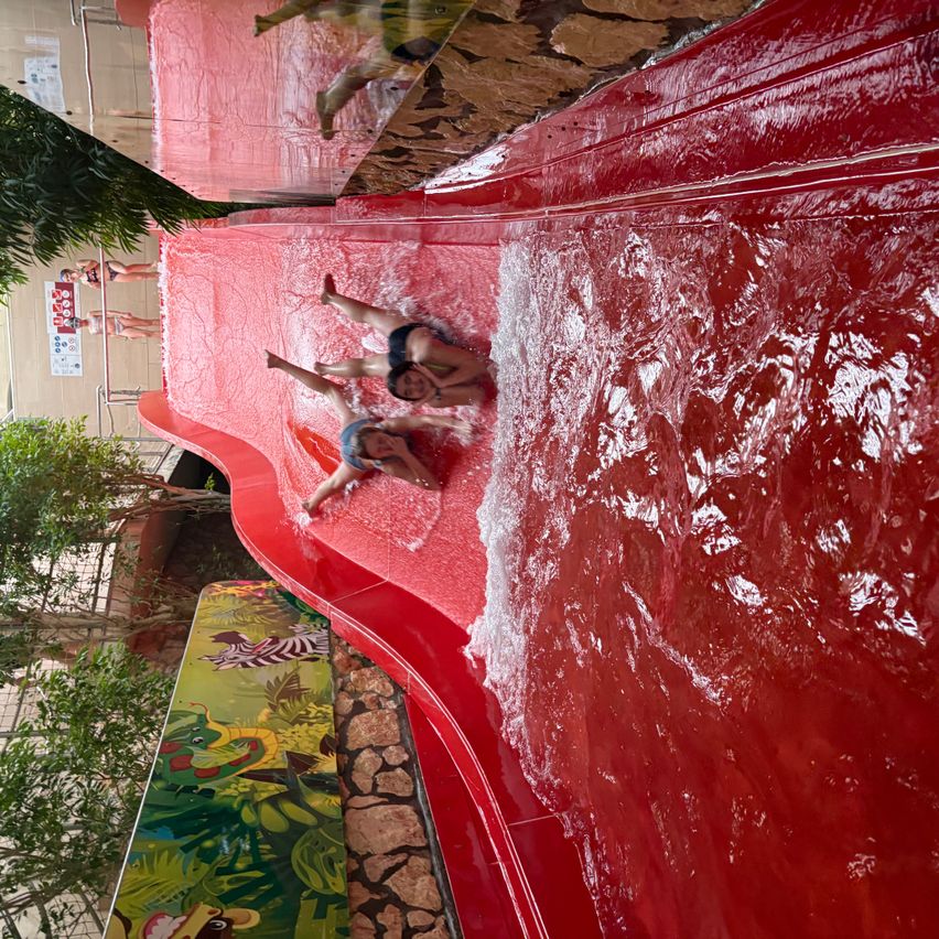 A view from above of people sliding down a red water slide. Water splashes around them as they descend. Trees and plants are visible in the background.