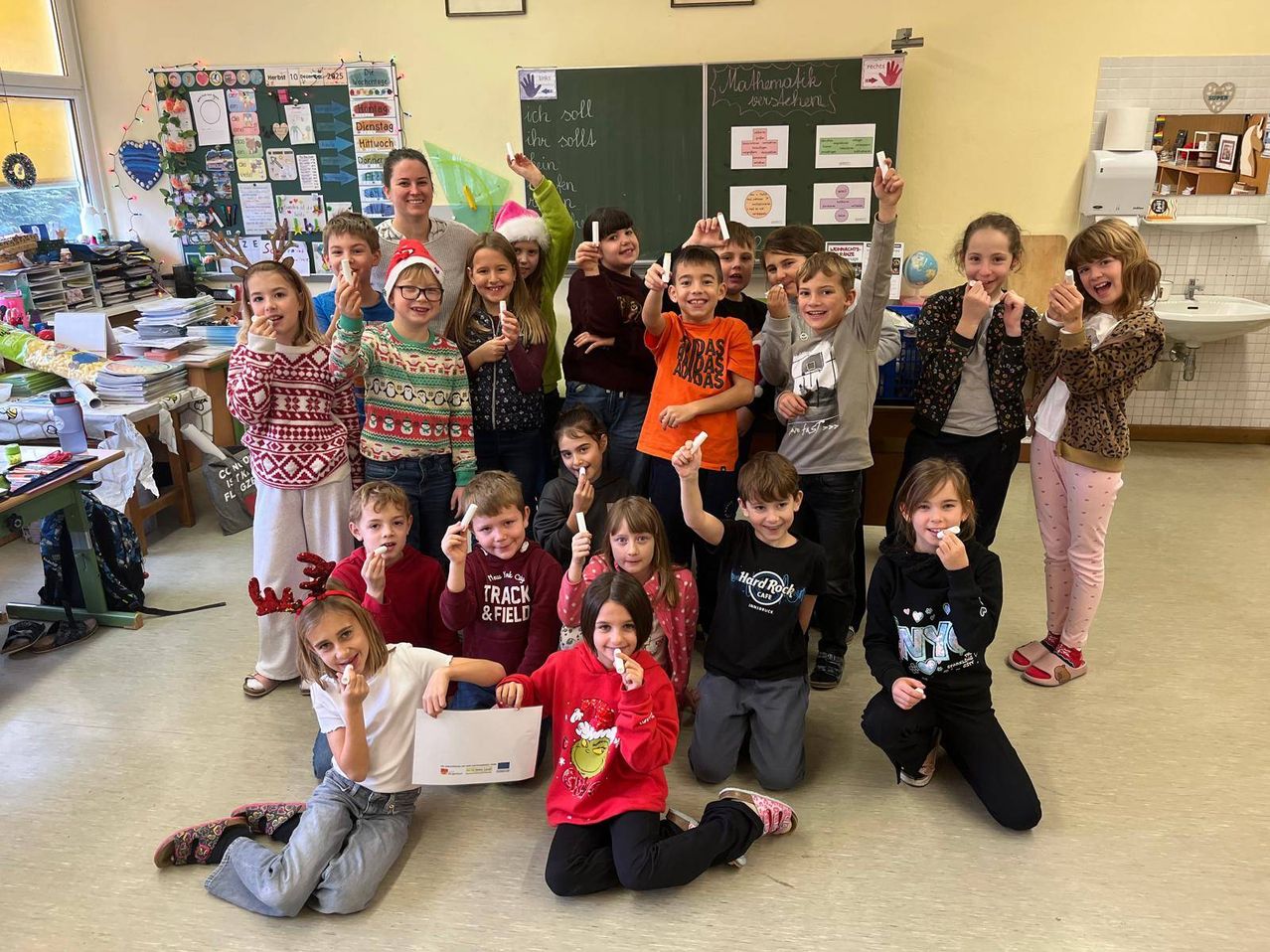 Ein Klassenzimmer voller Kinder, einige sitzend und einige stehend, halten Zahnbürsten und lächeln. Eine Lehrerin steht in der Mitte. Verschiedene Poster und Dekorationen sind an den Wänden.