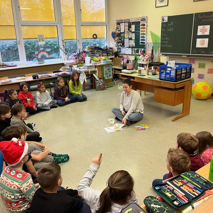 Ein Klassenzimmer mit Schülern, die auf dem Boden sitzen, während eine Lehrerin ihnen ein Buch vorliest. Das Klassenzimmer ist mit Postern, einem Regenbogen und einem großen Ball dekoriert.