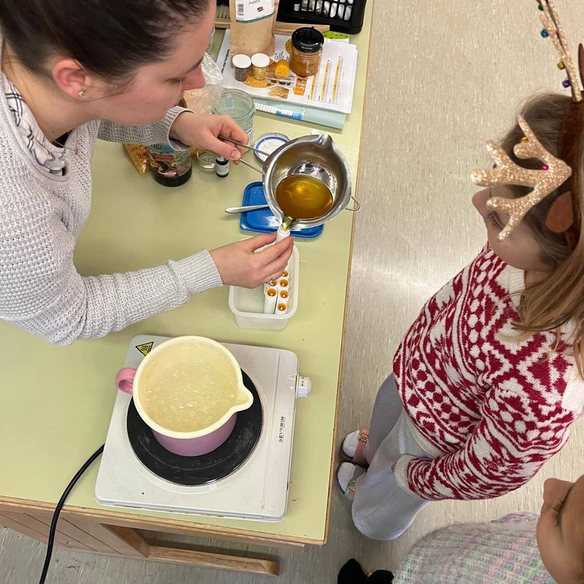 Eine Frau mit einer Halskette gießt Honig in eine Schüssel, während ein Kind mit einem Rehkitz-Kopfschmuck zusieht. Der Tisch hat einen Becher, Flaschen und Gläser.