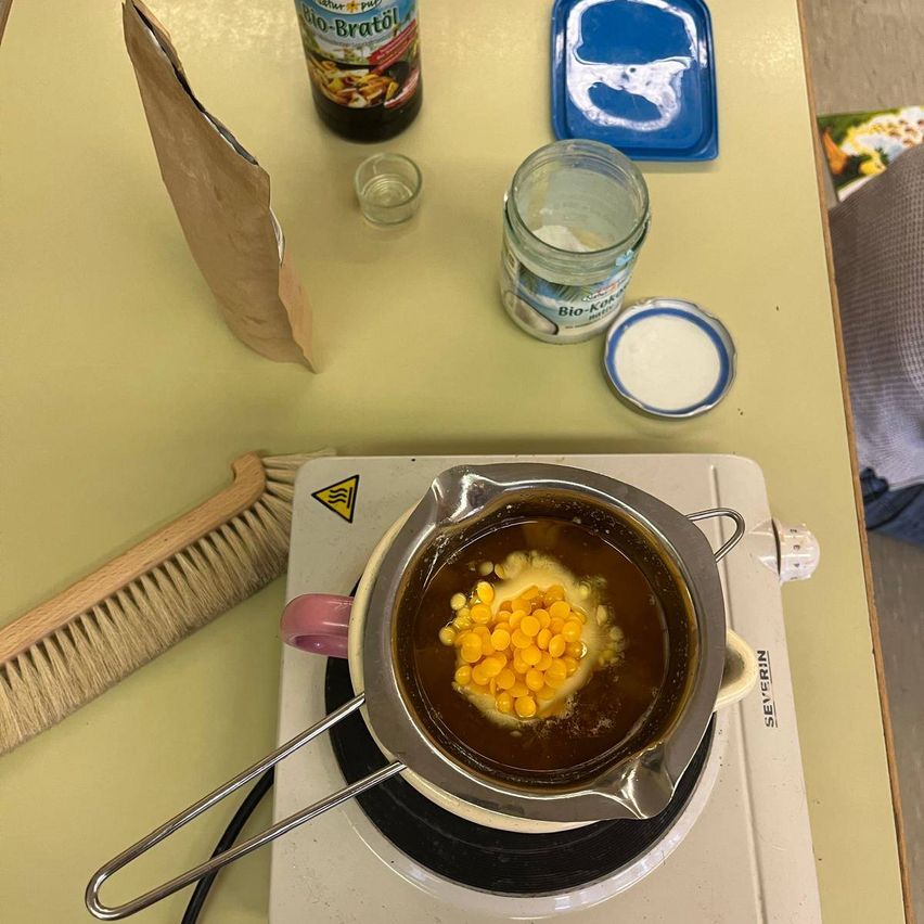 Ein Herd mit einem Topf darauf steht auf einem gelben Tisch. Der Topf hat eine gelbe Flüssigkeit und Maiskörner. In der Nähe sind eine Flasche, ein Becher, ein Glas und ein blauer Plastikbehälter. Ein Holzpinsel befindet sich links.