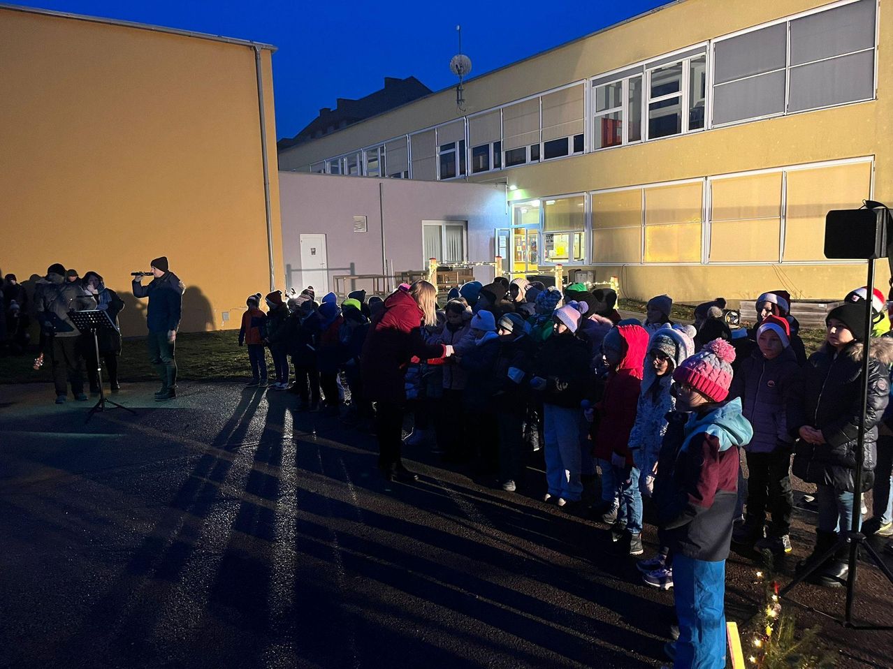 Eine Gruppe von Kindern steht vor einem Gebäude und trägt Winterkleidung und Mützen. Ein Mann spielt Flöte.