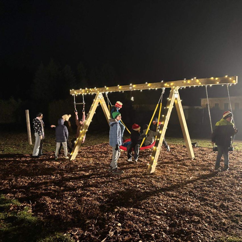Eine Gruppe von Kindern spielt auf einer mit Lichtern geschmückten Holzschaukel in einem Park bei Nacht.