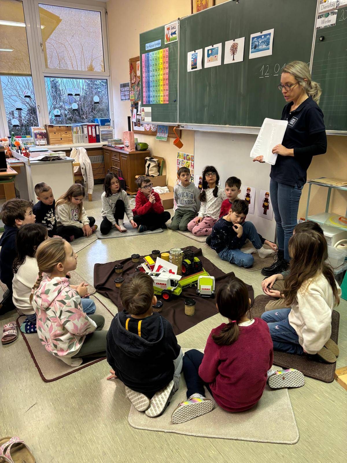 A woman stands in a classroom holding a piece of paper while young children sit in a circle listening. A brown blanket is on the floor with toys and snacks.