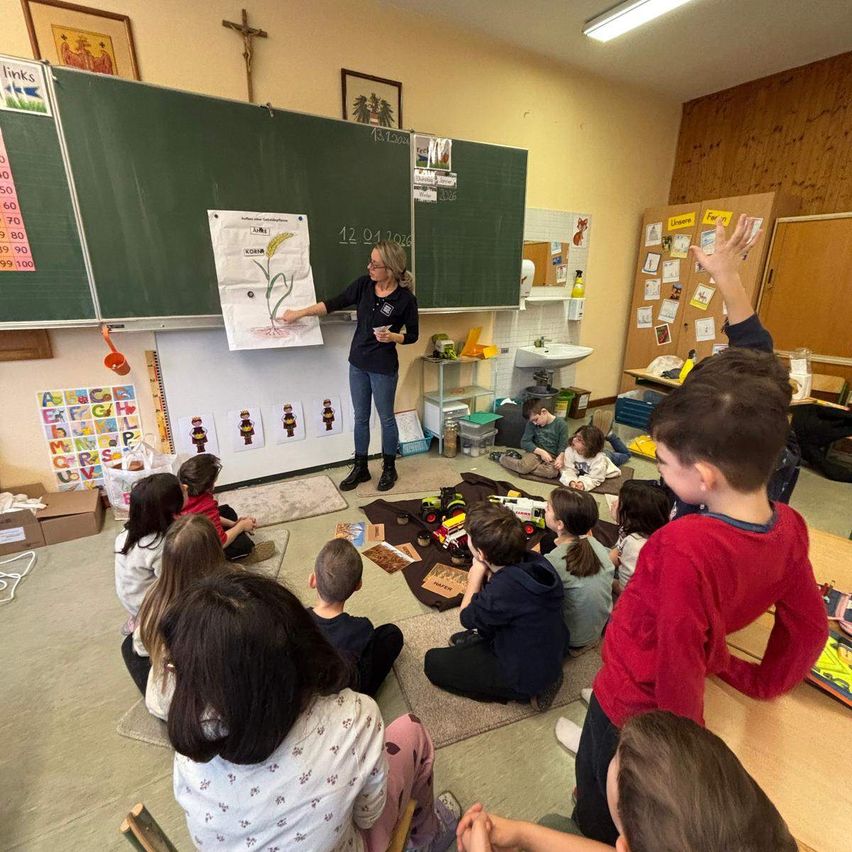 A teacher is teaching a classroom of children. The teacher is standing and holding a paper, showing something to the students. The classroom has a green board and a bulletin board. The students are sitting on the floor.