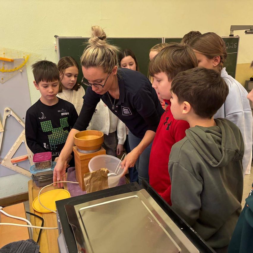 A woman in a black shirt and glasses is demonstrating something to a group of children in a classroom. They are looking at a machine with a bowl and a container on top.