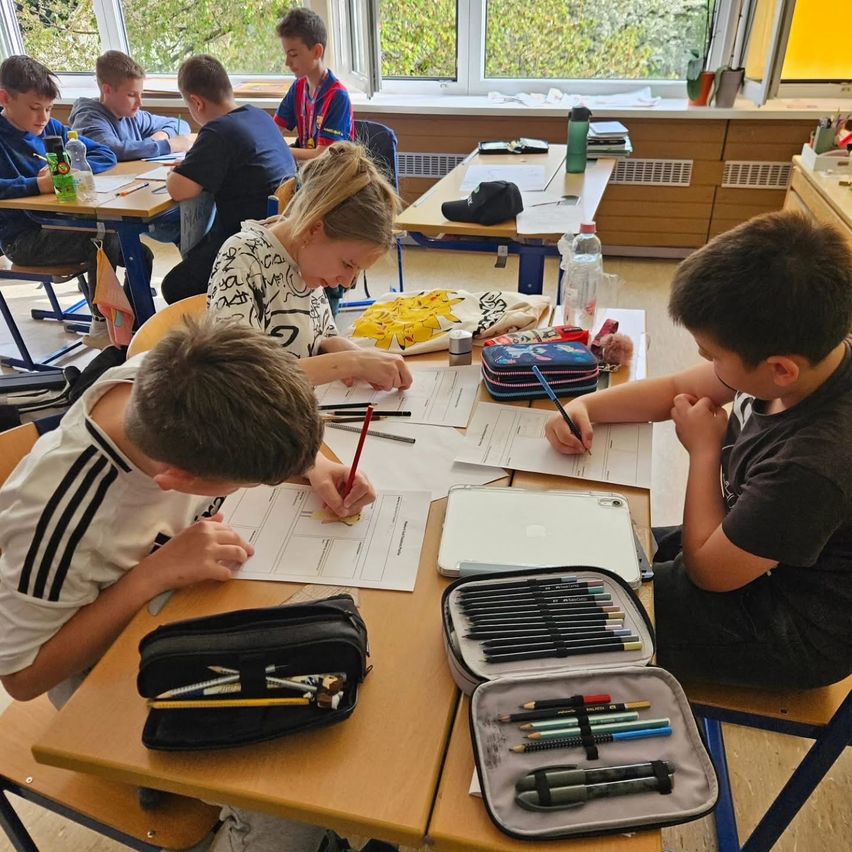 A classroom filled with young students, engaged in drawing activities, surrounded by desks and supplies, with trees visible outside the window.