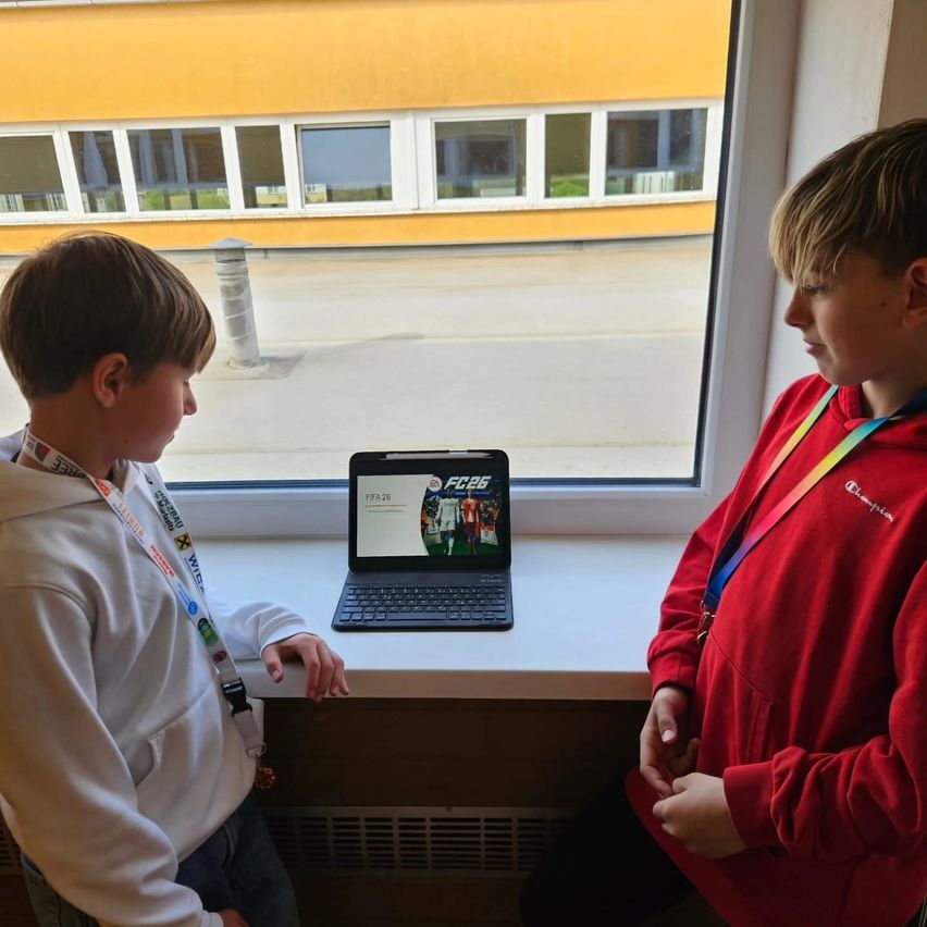Two boys are looking at a laptop with a soccer team on the screen. They are standing in front of a window with a view of a building and a road.