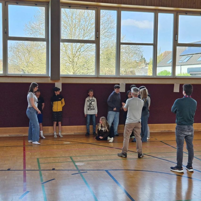 A group of people are standing and sitting in a gymnasium with large windows. The floor has blue and green markings.