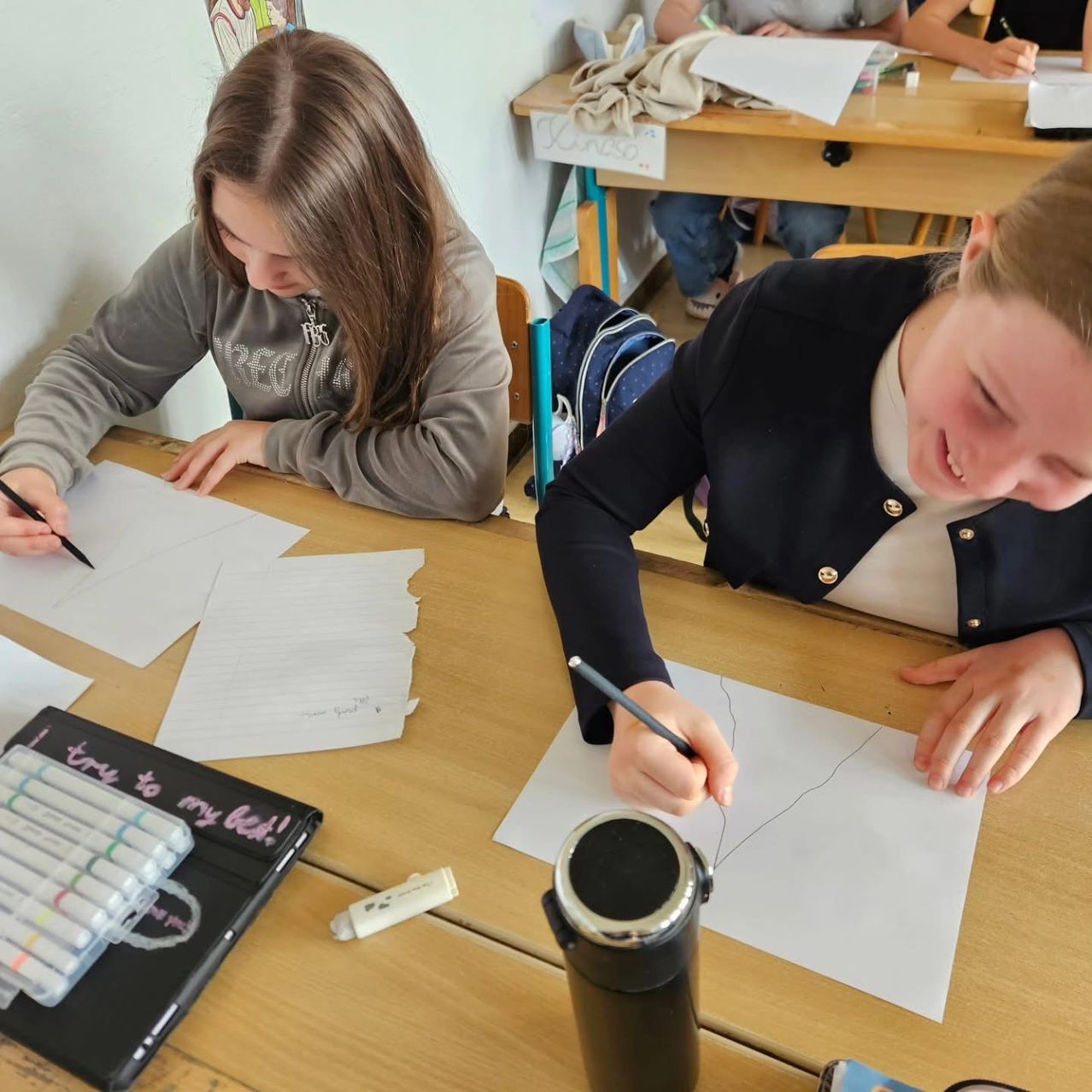 Two girls in a classroom, one focused on her work, the other smiling while drawing on paper. Both have papers and markers on their desks.