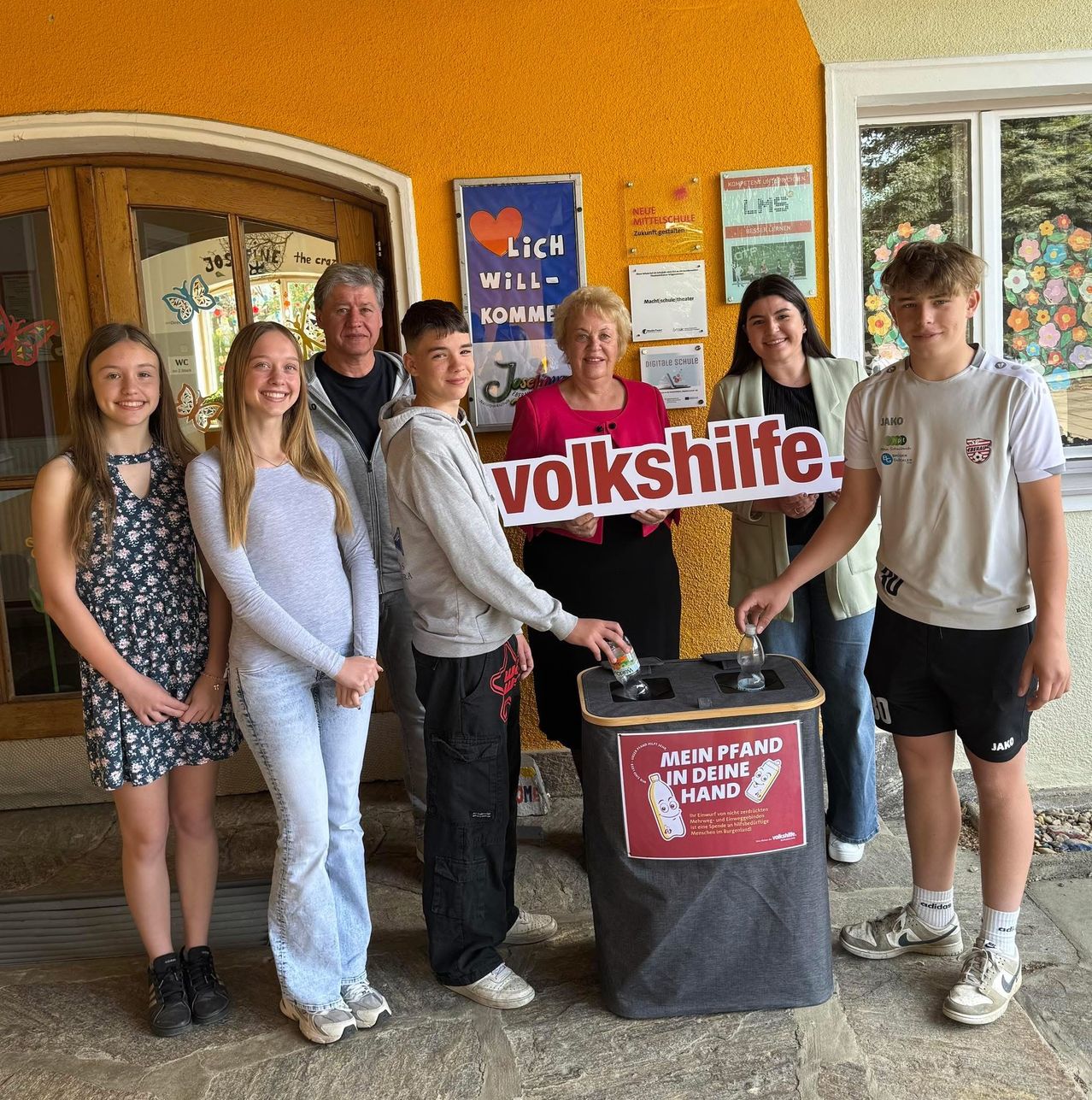 Eine Gruppe von Menschen steht neben einem Recycling-Behälter mit einem Schild, auf dem 'Volkshilfe' steht. Sie halten ein Schild, auf dem 'Mein Pfand in deine Hand' steht. Die Gruppe besteht aus einem Mann, einer Frau und vier jungen Leuten.
