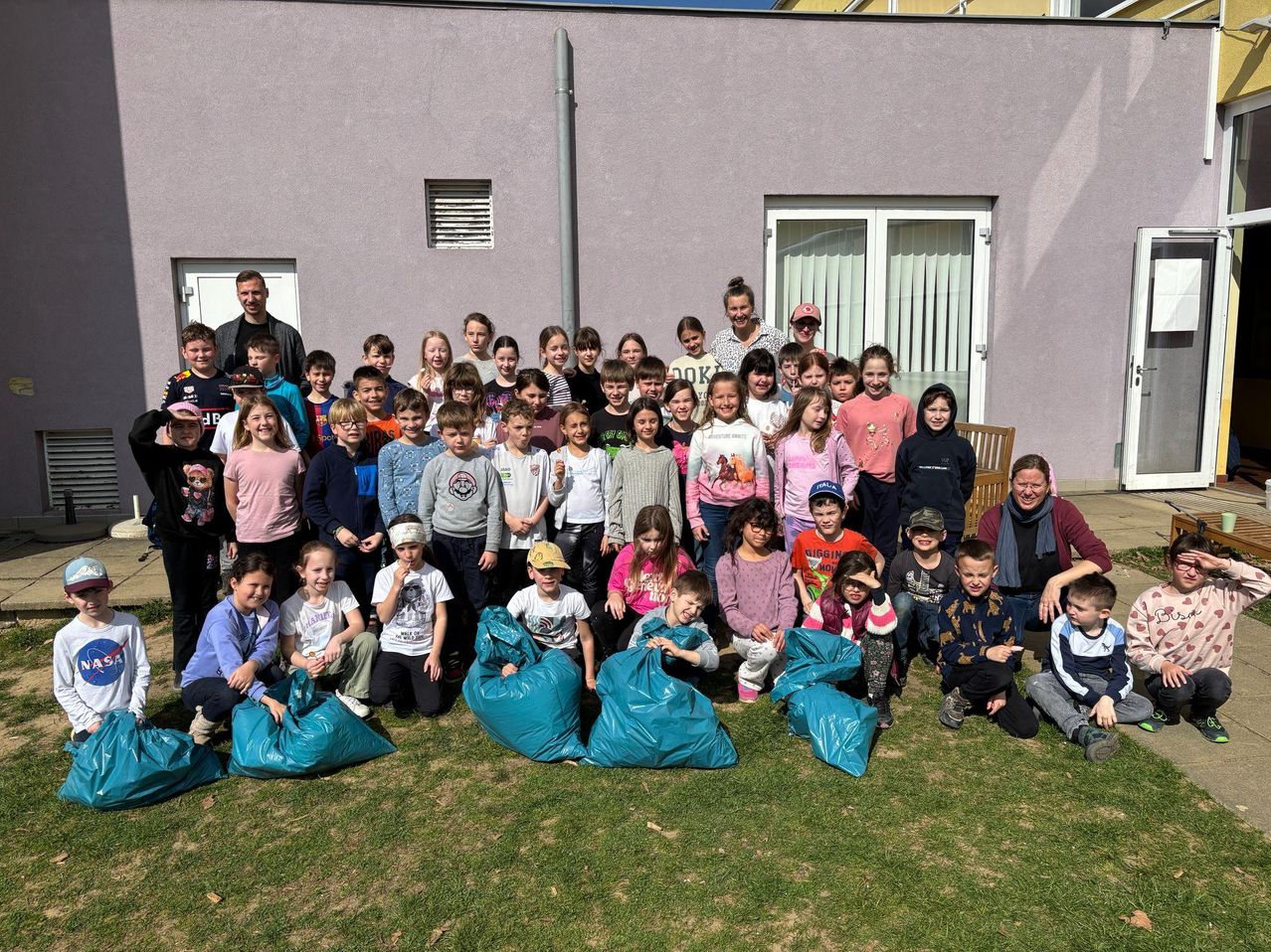 Eine Gruppe von Kindern und Erwachsenen posiert auf einem Rasen mit blauen Tüten vor ihnen für ein Foto. Ein Gebäude mit weißen Türen und Fenstern steht hinter ihnen.