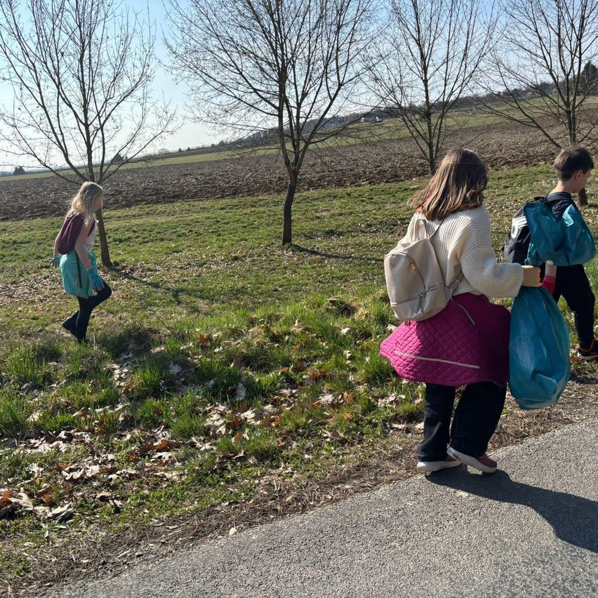 Zwei Kinder gehen neben einem Feld auf einem grasbewachsenen Pfad, wobei eines eine blaue Tasche und das andere einen Rucksack trägt. Im Hintergrund sind blattlose Bäume und eine entfernte Farm zu sehen.