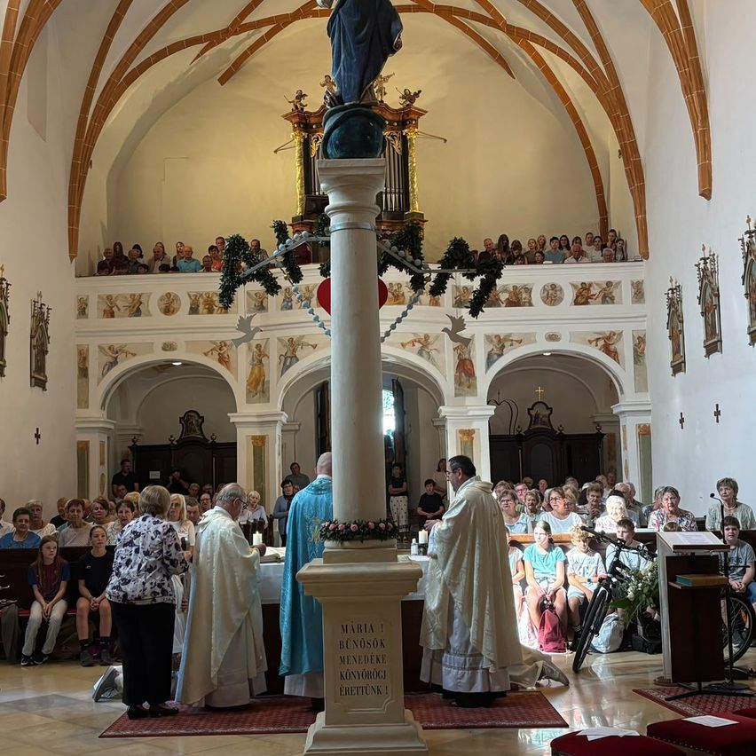 Priests conduct a ceremony in a church with a statue of Mary, surrounded by a congregation and decorative elements.
