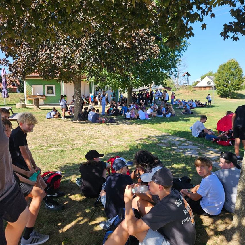 A group of people are sitting on the grass in a park, some under trees. Some are wearing hats and sneakers. A building is in the background.