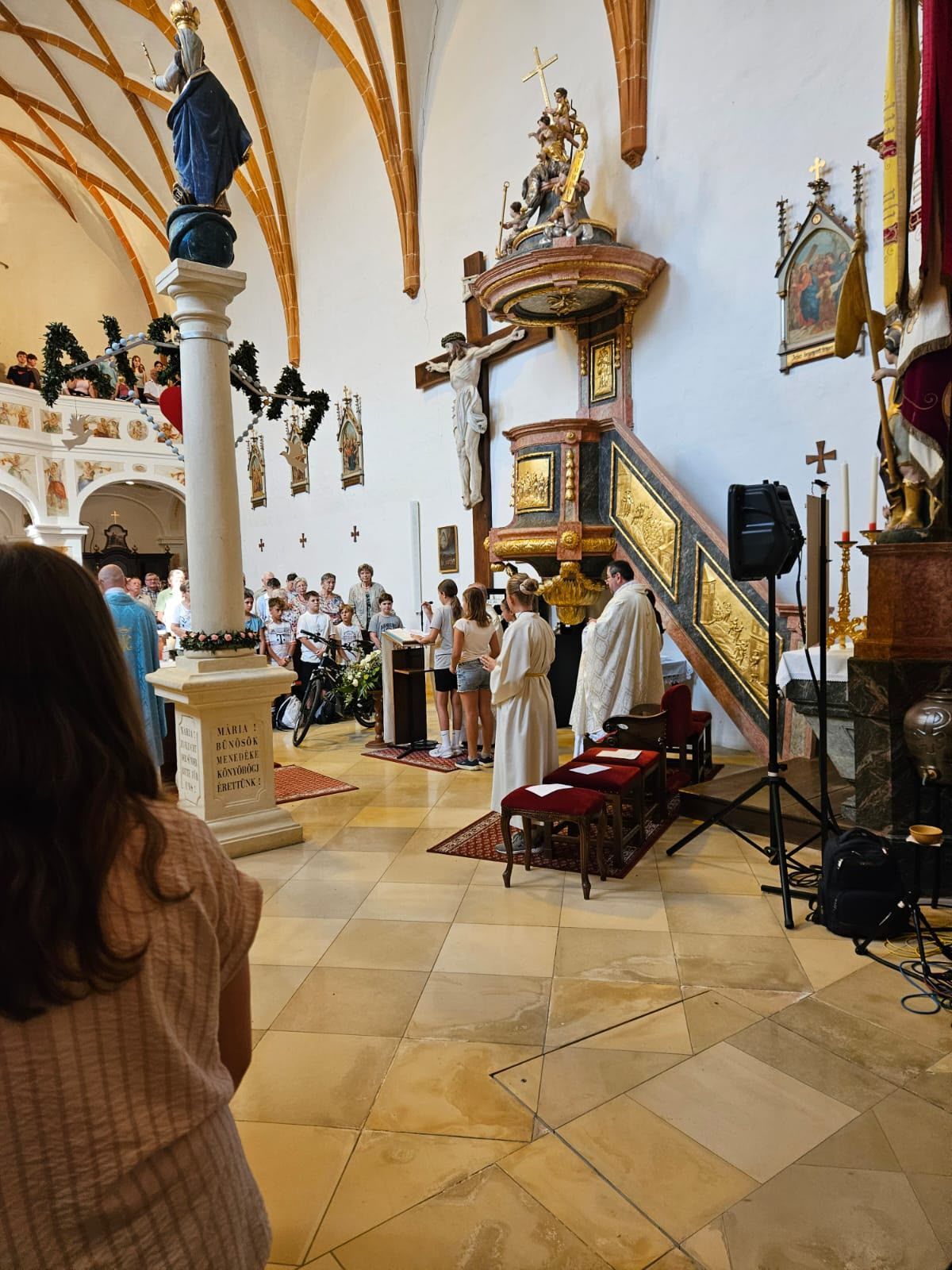 A group of people are gathered in a church, with a priest standing at the altar. A cross is mounted on the wall, and a statue is on the right. A few people are standing in front of the altar, and a few are sitting on the chairs. A speaker is placed on the tripod stand.
