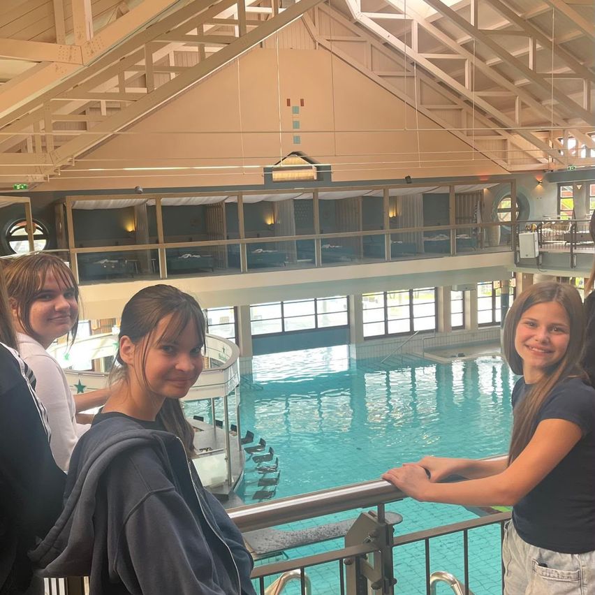Four girls stand by the railing of an indoor pool, smiling at the camera. The pool is large and filled with clear water. Above, a wooden ceiling with a triangular design.