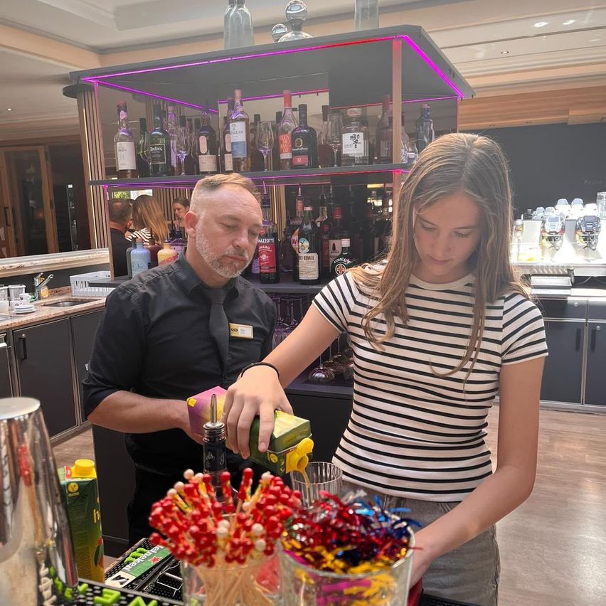 A bartender and a young girl are preparing drinks at a bar counter. The bartender is holding a bottle, and the girl is holding a box. There are colorful candies and a shaker on the counter. Behind them, there is a shelf with bottles of alcohol.